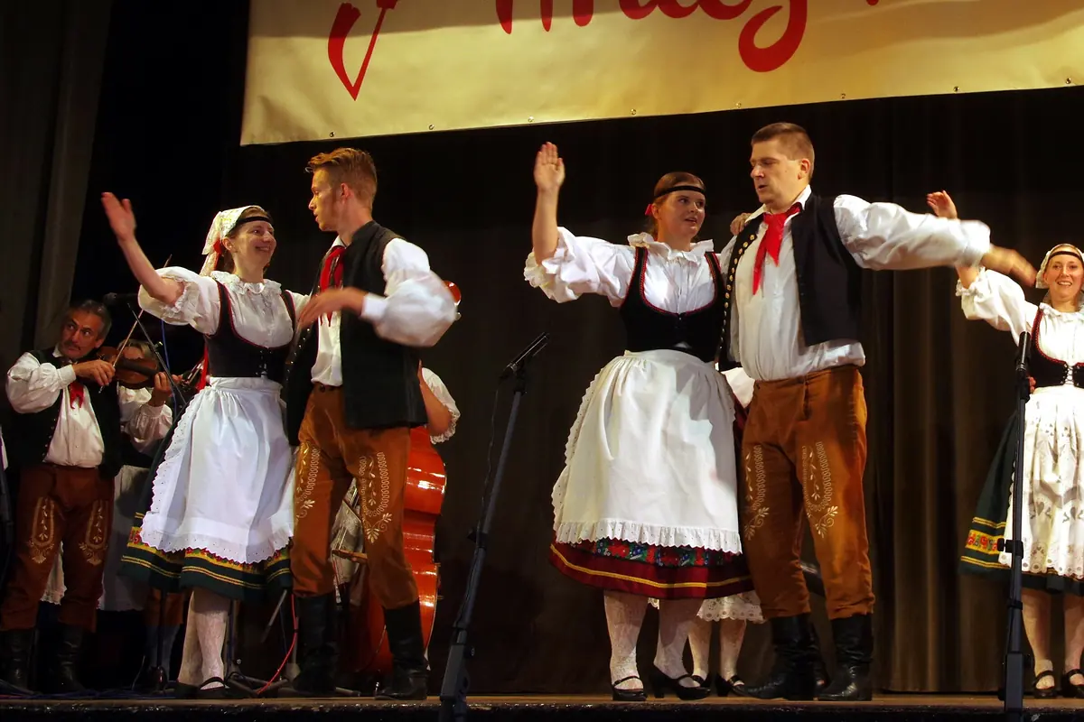 Adult folk dancers on a stage in costume, women in white aprons and black bodices, men in dark waistcoats, red neckerchiefs and brown trousers