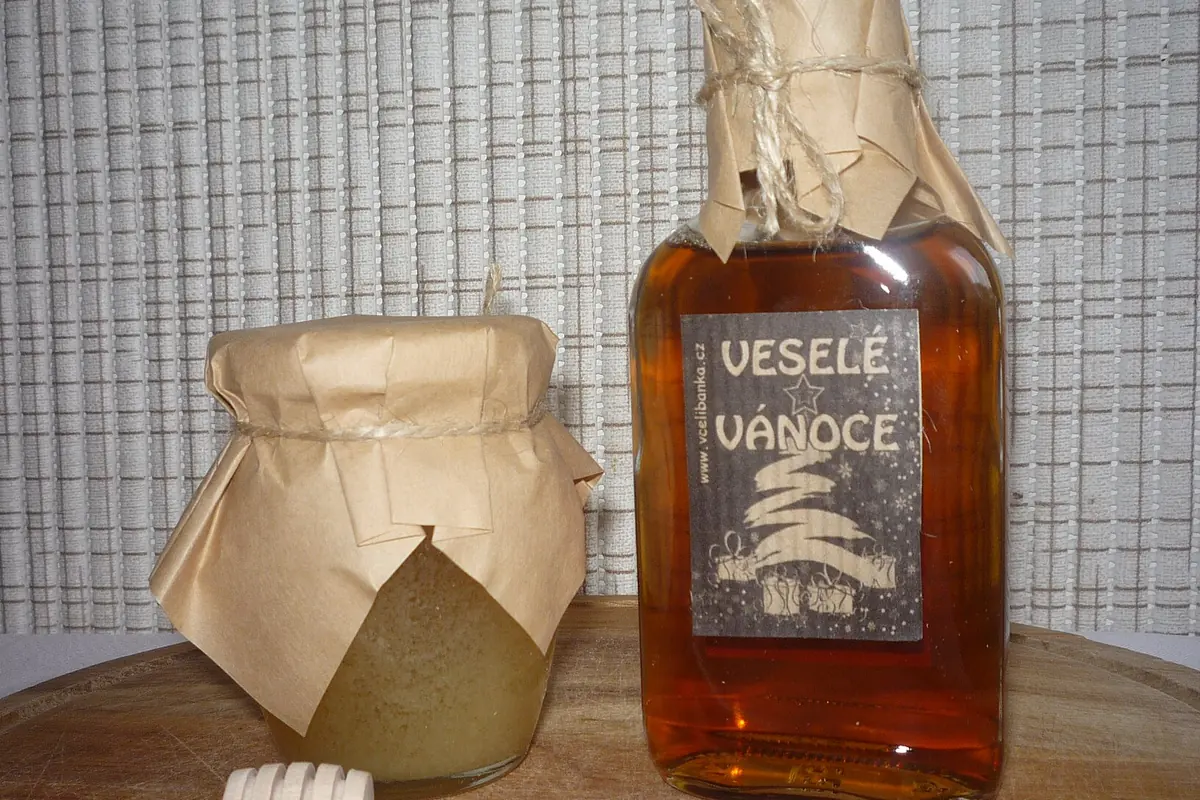 A jar of honey with a wooden dipper beside a glass bottle of amber medovina, the Czech word for mead, on a wooden board